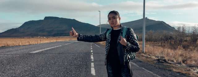 A woman wearing a leather jacket and a backpack standing by the side of the road with her thumb out. In the background