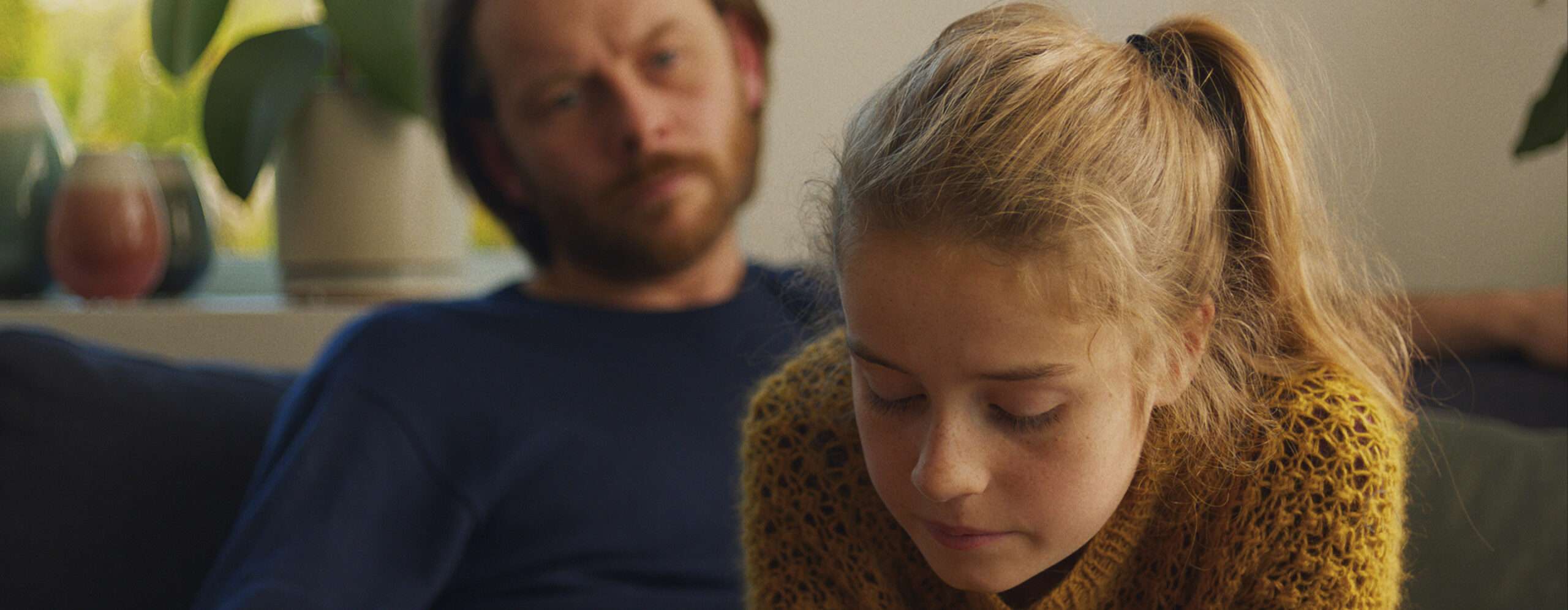 photo of a young blonde girl, behind her out of focus a man looking at her with a serious expression